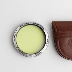 May include: A silver filter with a yellow-green glass lens in a brown leather case. The filter is labeled "Rollei - gelb - hell 285 ø" and "GERMANY FRANKE & HEIDECKE". The case has the Rollei logo embossed on it.