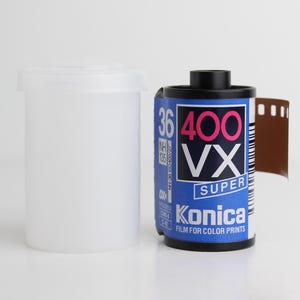 May include: A roll of Konica VX Super 400 film for color prints, with a blue and white label, next to a white plastic film canister. The film roll is black and has a brown film strip partially unrolled.