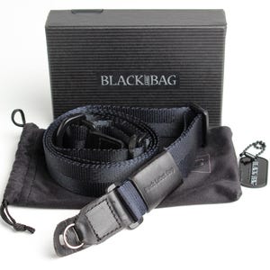 May include: Black camera strap with a black leather pad and a black bag with the text "Black  Bag" on it.