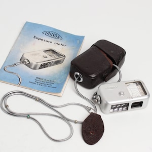May include: A silver Minox exposure meter with a black leather case and a silver chain. The meter has a small window with a scale and a button on the side. The case has a flap that closes with a snap. The chain has a small brown leather tag.