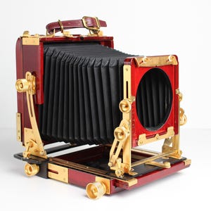 May include: A vintage-style large format camera with a red wooden body, gold-tone metal accents, and a black bellows.