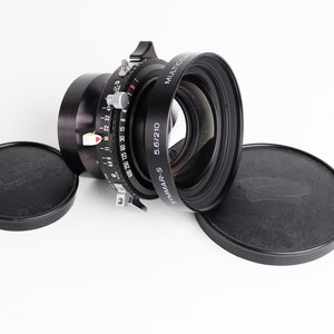 Op de afbeelding: Een zwarte cameralens met de tekst "SYMMAR-S 5.6/210" en "MULTICOAT" wordt weergegeven op een witte achtergrond. Twee zwarte lensdoppen bevinden zich aan weerszijden van de lens. De lens heeft verschillende meetmarkeringen.