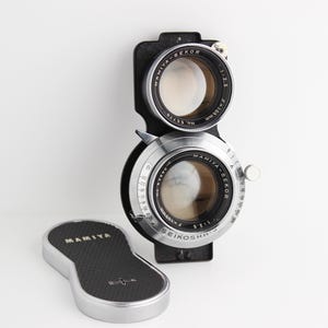 May include: A vintage Mamiya-Sekor camera lens with a black and silver design. The lens has two circular openings with the text "MAMIYA-SEKOR" and "f=105mm" visible. A silver lens cap with the "MAMIYA" logo is also present.