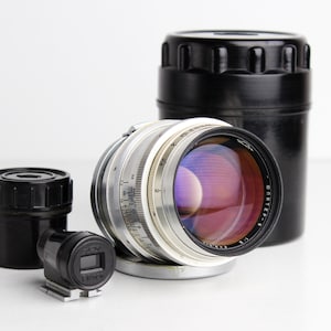 May include: A silver camera lens with a black lens cap and a black case. The lens has a silver ring with markings and the text "Jupiter-8" printed on it.