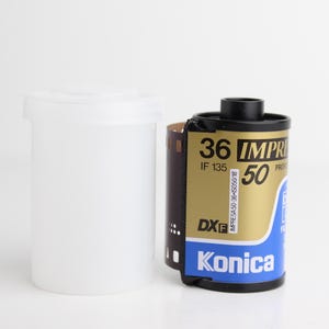May include: A roll of Konica film with a black cap, gold and blue label, and the text "36 IMPRESA 50". A white plastic film canister is next to the film roll. The film roll is partially unrolled.