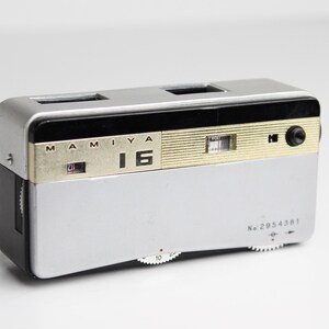 May include: A silver and black Mamiya 16 camera with gold trim and a black dial. The camera has a serial number "No. 2954381" printed on the bottom.
