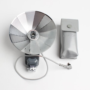 May include: A silver and gray flash unit with a Honeywell Flash-A-Matic label. The flash unit has a round reflector and a cord with a connector. It comes with a gray carrying case with the Honeywell logo.