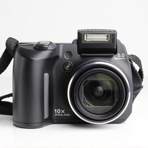 May include: A black Olympus digital camera with a 10x optical zoom and a 6.0 megapixel resolution. The camera has a black strap and a built-in flash.