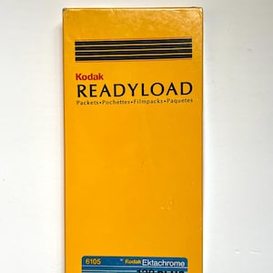 May include: A rectangular Kodak film box in bright yellow with black horizontal stripes. The box reads "READYLOAD" in large, bold letters, with smaller text below. The bottom has a blue and red stripe with "Kodak Ektachrome 100 PLUS" printed on it.