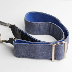 Puede incluir: Una correa de cámara azul y negra con una hebilla plateada. La correa está hecha de tejido tejido y tiene una sección de cuero con una cadena de metal.
