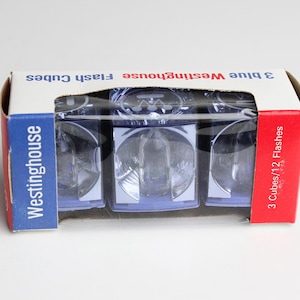 May include: A package of three blue Westinghouse flash cubes. The box is white with blue and red accents, and the text "3 blue Westinghouse Flash Cubes" is printed on the top. The side of the box reads "3 Cubes/12 Flashes".