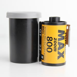 Può includere: Un contenitore per pellicola nero con coperchio grigio accanto a un rotolo di pellicola Kodak Max Zoom 800 da 35 mm. Il rotolo di pellicola è giallo con testo nero, tra cui "MAX Zoom 800". La pellicola è per stampe a colori.