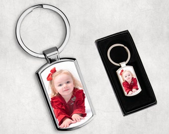 May include: A silver keychain with a rectangular photo insert featuring a young girl wearing a red dress and a bow in her hair. The keychain is in a box with a black velvet lining.