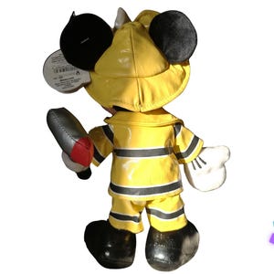 Puede incluir: Una figura de peluche de Mickey Mouse vestido de bombero, con abrigo, pantal&oacute;n y casco amarillos. La figura sostiene una herramienta de bombero plateada y roja. Las orejas y los zapatos negros completan el look. Una etiqueta est&aacute; adjunta.