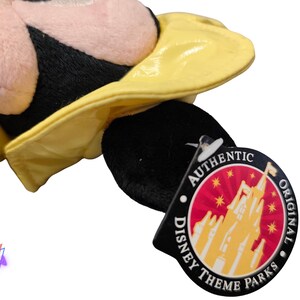 Puede incluir: Un peluche con cabeza negra y sombrero amarillo. El juguete tiene una cara rosa y es parcialmente visible. Una etiqueta negra con las palabras "AUTHENTIC DISNEY THEME PARKS ORIGINAL" est&aacute; adjunta.