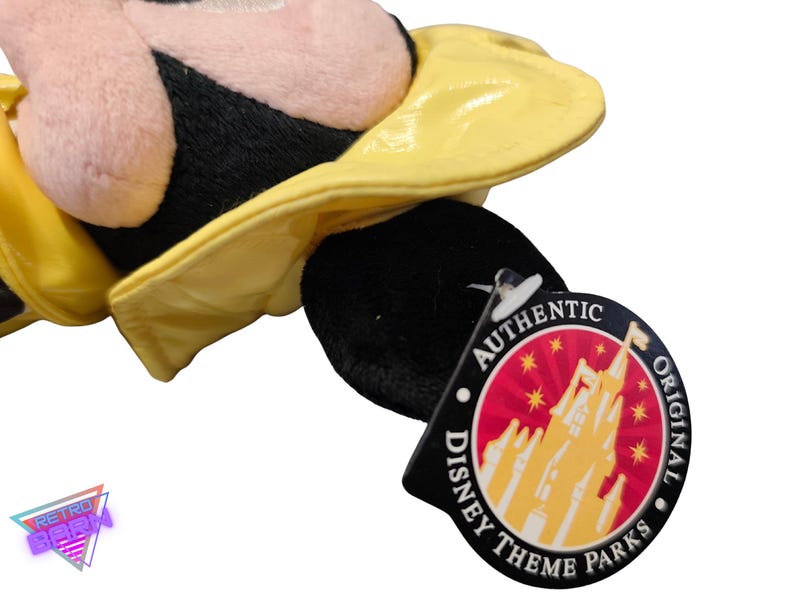 Puede incluir: Un peluche con cabeza negra y sombrero amarillo. El juguete tiene una cara rosa y es parcialmente visible. Una etiqueta negra con las palabras "AUTHENTIC DISNEY THEME PARKS ORIGINAL" est&aacute; adjunta.