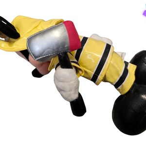 Puede incluir: Un peluche de un personaje de dibujos animados disfrazado de bombero. El juguete lleva una chaqueta amarilla con ribetes negros, guantes negros y botas negras. Sostiene un martillo de juguete plateado y rojo. Las orejas del personaje son negras.