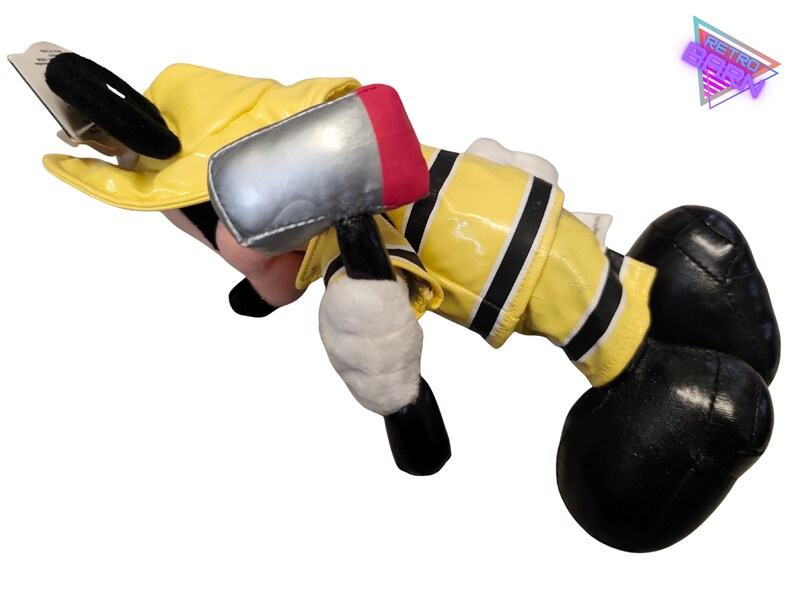 Puede incluir: Un peluche de un personaje de dibujos animados disfrazado de bombero. El juguete lleva una chaqueta amarilla con ribetes negros, guantes negros y botas negras. Sostiene un martillo de juguete plateado y rojo. Las orejas del personaje son negras.