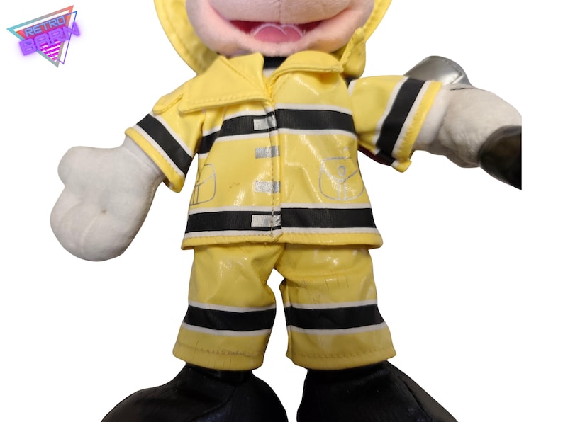 Puede incluir: Un peluche de un personaje con un uniforme de bombero amarillo con rayas negras. El juguete tiene guantes blancos y una cara sonriente. El uniforme incluye una chaqueta, pantalones y un casco.