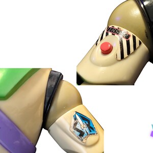 May include: Close-up of a Buzz Lightyear action figure. The figure features a cream-colored body with green, purple, and black accents. A red button and a sticker with the word "LASER" are visible. A blue and white emblem is on the arm.