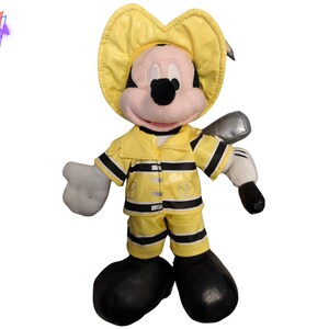 Puede incluir: Un peluche de Mickey Mouse vestido de bombero. El juguete lleva un casco, una chaqueta y un pantal&oacute;n de bombero amarillos con rayas negras. Tiene zapatos negros, guantes blancos y sostiene un hacha plateada. El juguete tiene una cara rosa y orejas negras.
