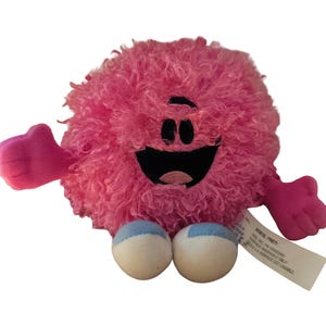 May include: A round, pink plush toy with a shaggy, textured surface. It features a black embroidered mouth and eyes, two pink arms with three fingers each, and two white feet with light blue accents. A white fabric tag is attached to the side.