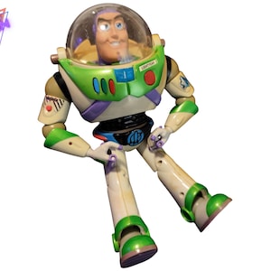 May include: A Buzz Lightyear action figure, featuring a clear helmet, green and white spacesuit, and a smiling face. The figure is in a seated position with its arms and legs extended. The figure has a "LIGHTYEAR" label on the helmet.