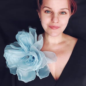 May include: A light blue fabric flower, possibly a brooch or shoulder accent, is pinned to a black top. The flower has multiple layers of delicate petals and a small, sparkling center. The woman in the image is wearing the flower.