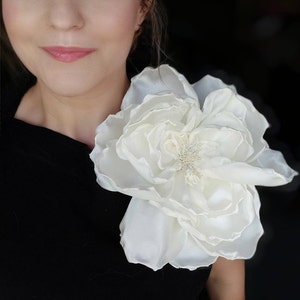 May include: A large, white, fabric flower brooch with a textured center. The flower is pinned to a black garment.