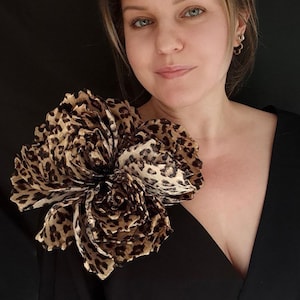 Op de afbeelding: Een grote stoffen bloem met een luipaardprint is vastgemaakt aan een zwart kledingstuk. De bloem is gemaakt van gelaagde, gerimpelde stof in tinten bruin, beige en crème. Het zwarte kledingstuk heeft een diepe V-hals.