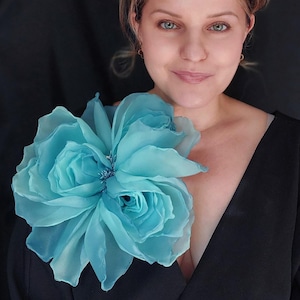 Può includere: Un grande fiore di tessuto azzurro, simile a una rosa, è appuntato su un capo nero. Il fiore ha più strati di petali delicati. Lo sfondo è nero pieno, creando un contrasto con il colore del fiore.