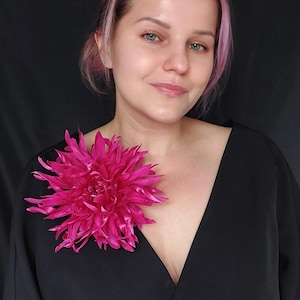 Peut inclure: Une grande broche en tissu de couleur rose vif. La fleur est une dahlia avec de nombreuses pétales et est portée sur une robe noire.