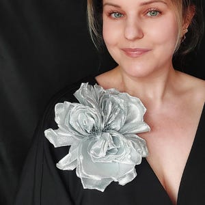 May include: A silver satin flower brooch with a layered design. The flower is a light silver colour and has a delicate, feminine look.