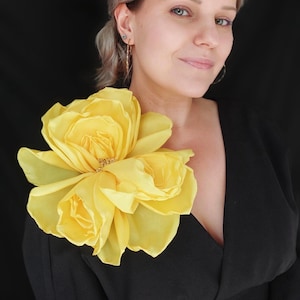 Könnte beinhalten: Eine große Brosche aus gelbem Stoffblumen, die auf der Schulter einer Person getragen wird, die ein schwarzes Kleid trägt.