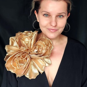 Könnte beinhalten: Eine große goldene Stoffblumenbrosche mit einem mehrschichtigen Design. Die Brosche wird auf einem schwarzen V-Ausschnitt-Oberteil getragen.