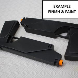 Puede incluir: Dos pistolas de juguete de plástico negro con puntas naranjas. Las pistolas están en estado de desmontaje, con el cañón y el mango de una pistola separados del cuerpo de la otra pistola. La imagen está etiquetada como "EXAMPLE FINISH & PAINT".