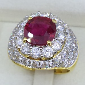 May include: A gold ring with a large, red, oval-shaped gemstone surrounded by a halo of smaller, white gemstones.