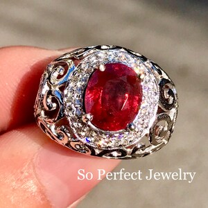 Könnte beinhalten: Ein silberner Ring mit einem großen ovalen Rubin, der von kleineren weißen Edelsteinen umgeben ist. Der Ring hat ein filigranes Design und der Text "So Perfect Jewelry" ist unterhalb des Rings sichtbar.