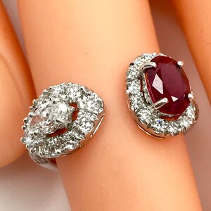 May include: A white gold ring with a large red ruby and smaller diamonds set in a halo around the ruby. The ring is open on one side and has a pear-shaped diamond on the other side.