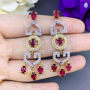 May include: A pair of gold earrings with dangling ruby and diamond accents. The earrings feature a geometric design with a circle in the center and three ruby drops hanging from the bottom.