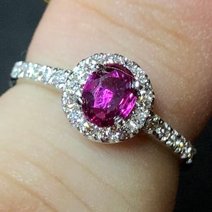 May include: A silver ring with a pink gemstone surrounded by a halo of smaller diamonds. The ring is set with a band of smaller diamonds.