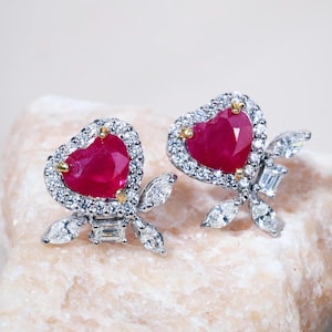 May include: A pair of stud earrings featuring a central heart-shaped red ruby surrounded by a halo of small round white stones. Below each heart, a cluster of marquise-cut and baguette-cut white stones forms a leaf-like accent in silver-tone metal.