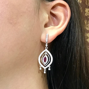May include: A pair of silver dangle earrings with a red gemstone in the center. The earrings are adorned with small clear stones.