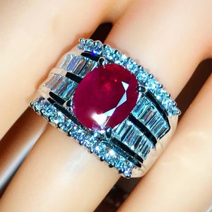 以下が含まれることがあります： 大きな楕円形のルビーを、小さなダイヤモンドのバンドで囲んだリング。リングはホワイトゴールド製です。