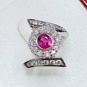 May include: A white gold ring with a pink gemstone surrounded by smaller diamonds. The ring has a unique design with two bands that intersect.