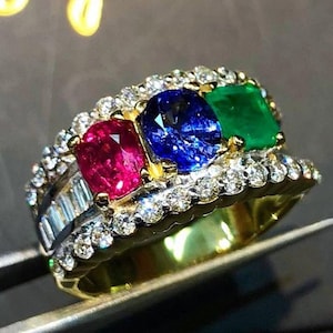 May include: A gold ring with a row of three gemstones: a red ruby, a blue sapphire, and a green emerald. The ring is set with smaller diamonds around the gemstones.