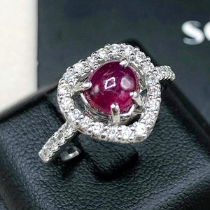 May include: A heart-shaped ring with a red ruby gemstone surrounded by a halo of smaller diamonds. The ring is set in white gold.
