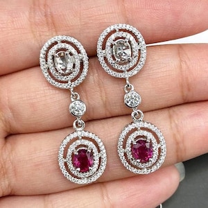May include: Dangling earrings with a circular design. Each earring features a central oval-shaped ruby, surrounded by a halo of small, sparkling white stones. The earrings are connected by a series of smaller, round white stones. The earrings are held in a hand.