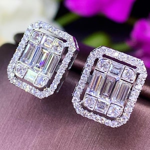 May include: A pair of silver earrings with a square-shaped design featuring a large, rectangular-cut diamond surrounded by smaller diamonds.