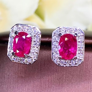 May include: A pair of ruby and diamond earrings. The earrings are set in a silver metal and feature a red oval ruby surrounded by a halo of smaller diamonds.
